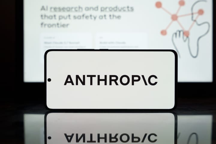 A mobile phone showing the Anthropic logo against a blurred background in which a computer screen can be seen